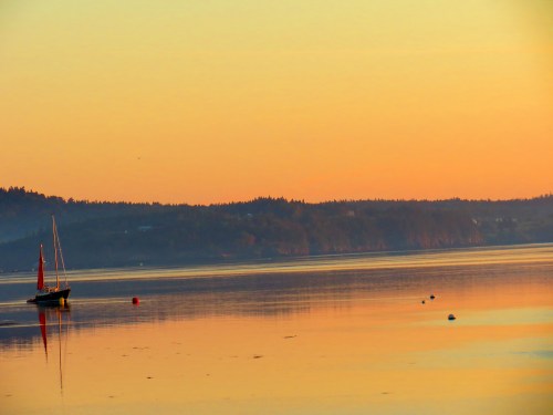 A calm bay at sunset, featuring a sailboat and reflections on the water, with hills in the background.