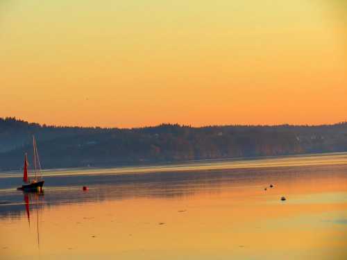 A calm bay at sunset, featuring a sailboat and reflections on the water, with hills in the background.