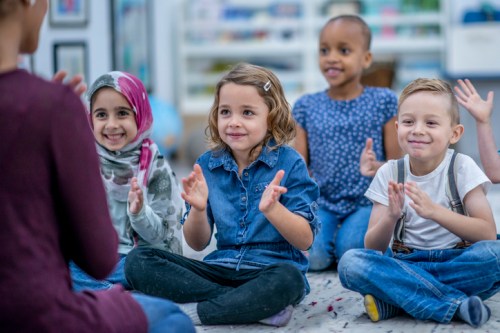 A group of four children sitting on the floor, clapping and smiling, engaged in a classroom activity.