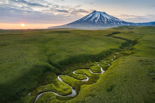 A winding river flows through lush green fields, with a snow-capped mountain rising in the background at sunset.