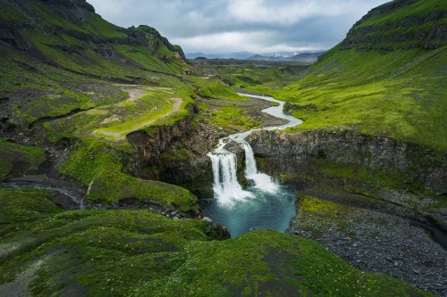 A scenic view of a waterfall cascading into a lush green valley, surrounded by rocky hills and overcast skies.