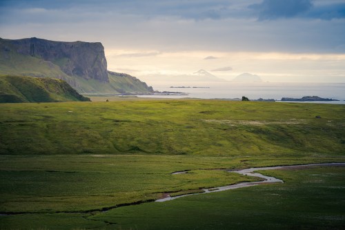 A serene landscape featuring green hills, a winding stream, and distant mountains under a cloudy sky by the coast.
