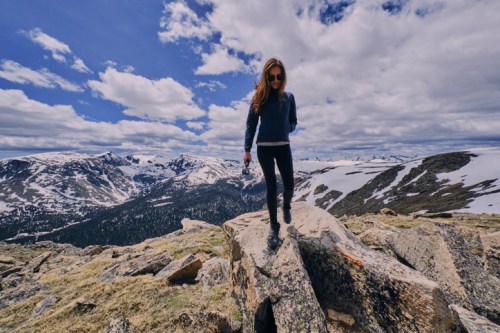 A woman walks on rocky terrain with a mountainous landscape and cloudy sky in the background.
