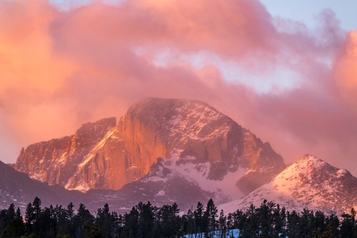 A majestic mountain peak bathed in warm sunset light, surrounded by soft pink clouds and silhouetted trees.