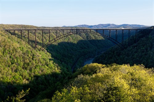 A tall, arched bridge spans a lush green valley, with rolling hills in the background under a clear blue sky.