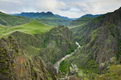 A lush green valley with a winding river, surrounded by steep rocky hills and distant mountains under a cloudy sky.