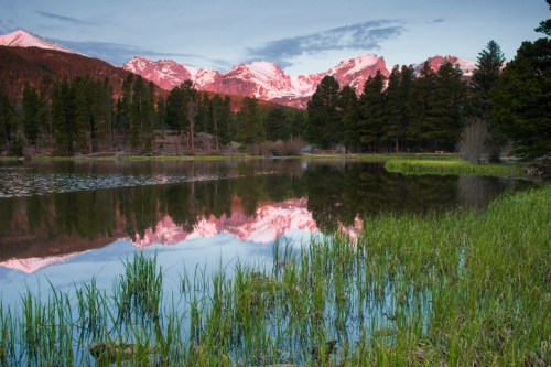 Serene lake reflecting snow-capped mountains and lush greenery at sunset, surrounded by tall trees.