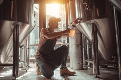 A person in a cap kneels by brewing tanks, pouring beer into a bottle with sunlight streaming through the window.