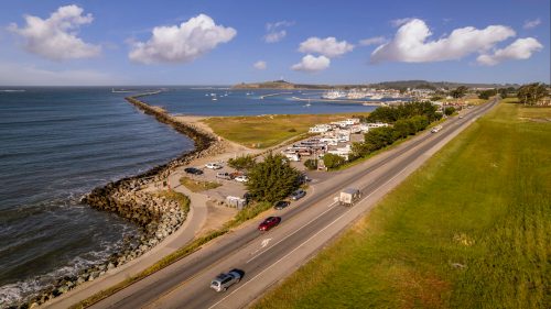 A coastal view featuring a road, parked RVs, and a harbor with boats under a blue sky with clouds.