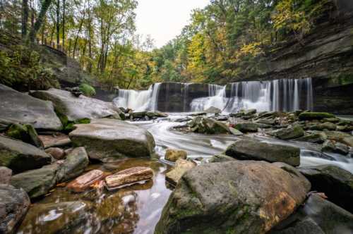 A serene waterfall cascades over rocks, surrounded by lush greenery and autumn foliage.