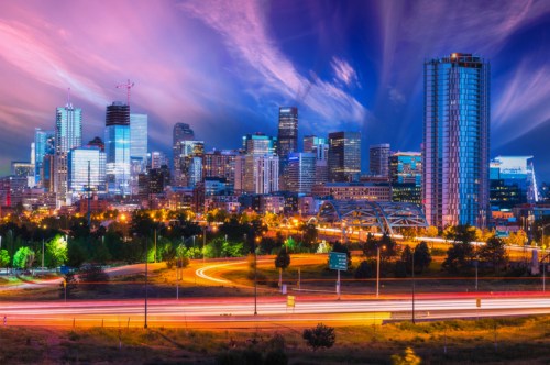 A vibrant city skyline at dusk, featuring tall buildings, colorful clouds, and light trails from passing cars.
