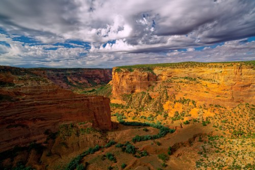 A scenic view of a canyon with orange cliffs, green vegetation, and a winding river under a cloudy sky.