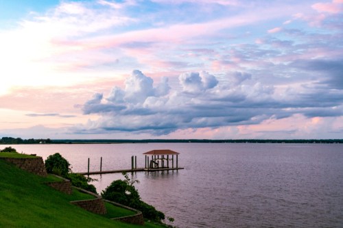 A serene lake at sunset, with colorful clouds and a small dock extending into the water. Green hills frame the scene.