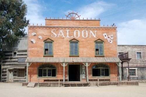 A wooden saloon building with "SALOON" sign, surrounded by a dusty western town under a blue sky.
