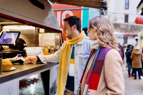 A man and woman in winter attire are ordering food at a food truck, with a bustling market scene in the background.