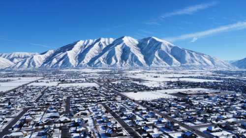 Aerial view of a snowy landscape with houses and a large mountain range in the background under a clear blue sky.