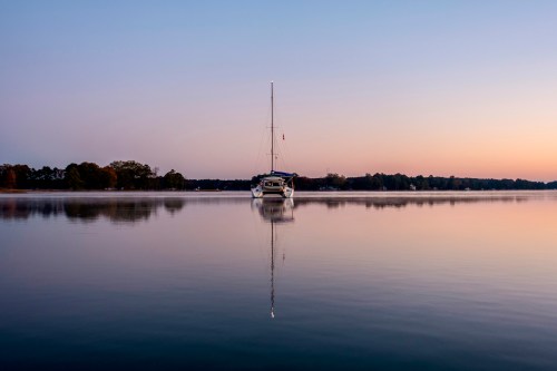 A calm lake at sunset with a sailboat anchored peacefully, reflecting on the water's surface.