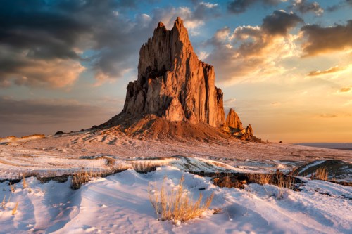 A dramatic rock formation rises from a snowy landscape under a colorful sunset sky.