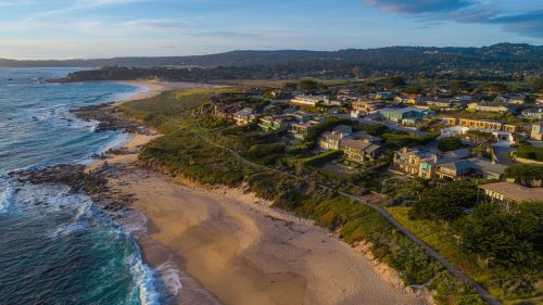Aerial view of coastal homes along a sandy beach, with waves crashing and hills in the background under a clear sky.