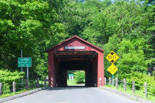 A red covered bridge with signs for Sharon and a warning for a one-lane bridge, surrounded by lush greenery.