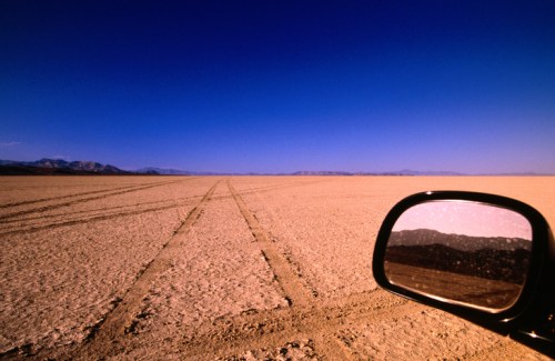 A vast, dry landscape with tire tracks leading into the distance, reflecting blue sky and mountains in a side mirror.