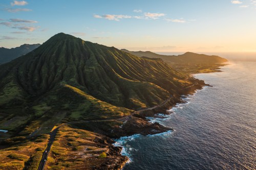 Aerial view of a lush green mountain overlooking the ocean at sunset, with a winding road along the coastline.