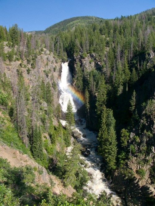 A waterfall cascades down rocky cliffs, surrounded by lush green trees and a rainbow forming in the mist.