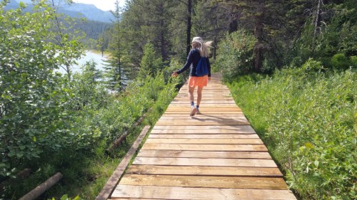 A person walks along a wooden path surrounded by greenery and trees near a lake.