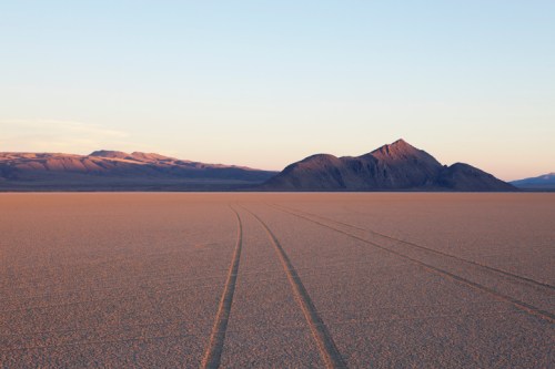 A vast desert landscape with tire tracks leading towards a distant mountain under a clear sky at sunset.