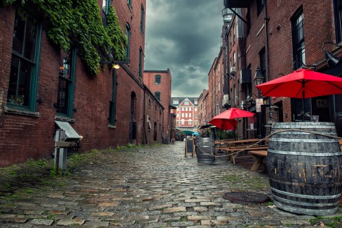 A cobblestone alley lined with red umbrellas, brick buildings, and wooden barrels under a cloudy sky.