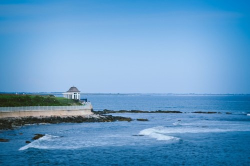 A serene coastal view featuring a gazebo on a rocky shore with gentle waves and a clear blue sky.