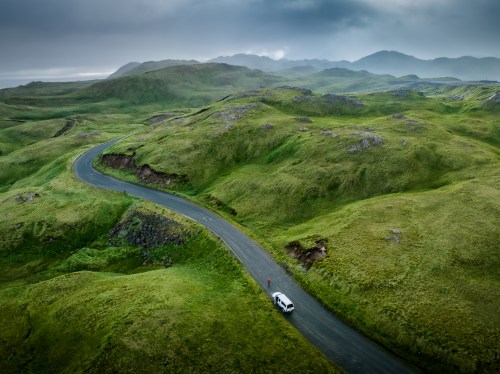 A winding road cuts through lush green hills under a cloudy sky, with a vehicle parked nearby.