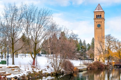 A clock tower beside a snowy park and calm water, with leafless trees and a blue sky in the background.