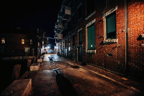 A dimly lit brick alley at night, with reflections on wet pavement and buildings lining the sides.