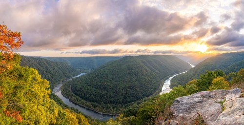 A panoramic view of a winding river surrounded by lush green hills under a cloudy sunset sky.