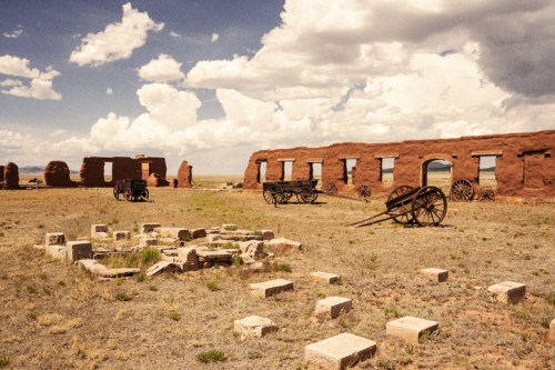 Ruins of a historic building with old wagons and stone remnants under a cloudy sky in a barren landscape.