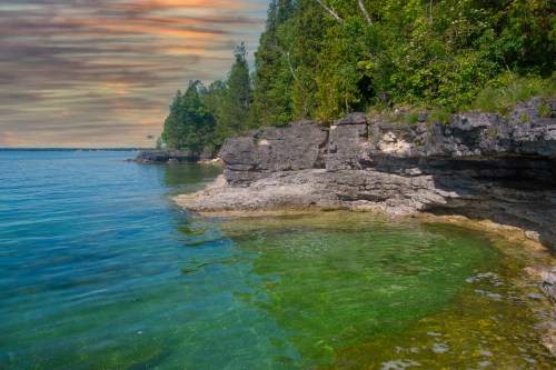 A serene shoreline with rocky cliffs and clear green water under a colorful sky at sunset.