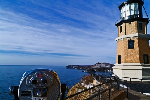 A lighthouse stands by the shore, with a scenic view of the ocean and a coin-operated viewer in the foreground.