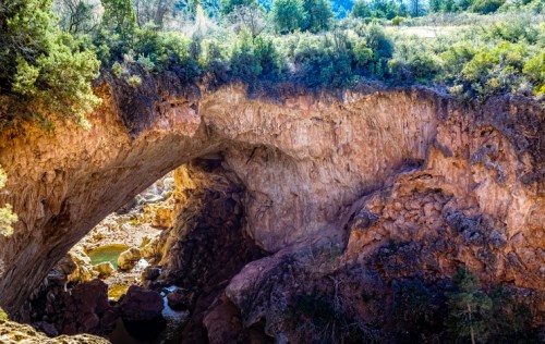 Natural rock arch over a river, surrounded by lush greenery and rocky terrain under a clear blue sky.