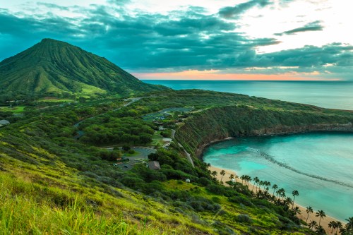A scenic view of a lush green coastline with a mountain, beach, and calm ocean under a cloudy sky at sunset.