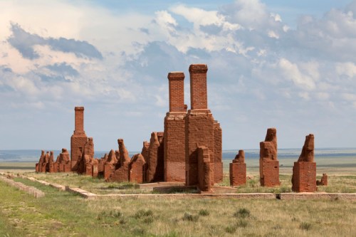 Ruins of brick structures with tall chimneys, set against a cloudy sky and grassy landscape.
