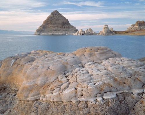 A rocky shoreline with textured formations in the foreground, leading to a calm lake and a distant pyramid-shaped island.