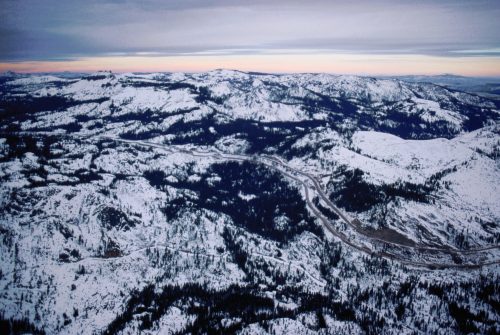 Aerial view of snow-covered mountains and a winding road under a cloudy sky at dusk.