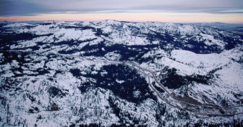 Aerial view of snow-covered mountains and winding roads under a soft, pastel sky at dusk.