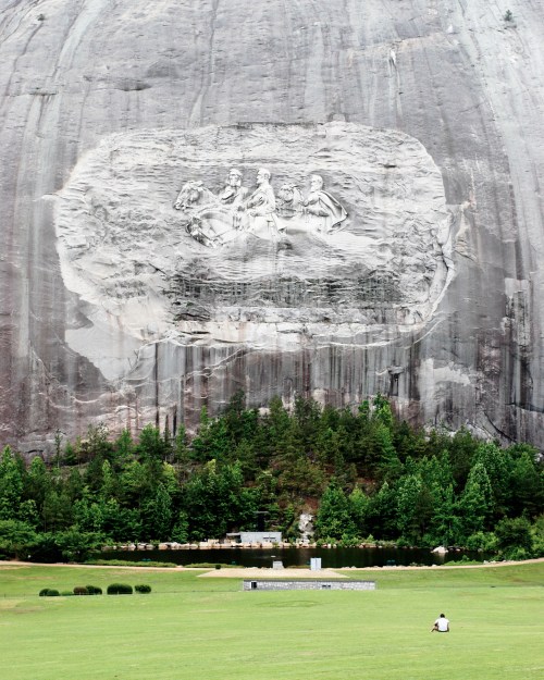 A large stone carving of three figures on a mountain, with a grassy area and trees in the foreground.