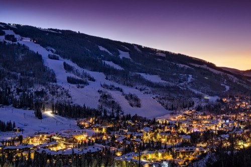 A snowy mountain village illuminated at dusk, with ski slopes and trees in the background under a twilight sky.