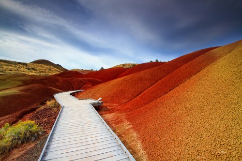 A wooden walkway winds through vibrant, multicolored hills under a blue sky, showcasing unique geological formations.