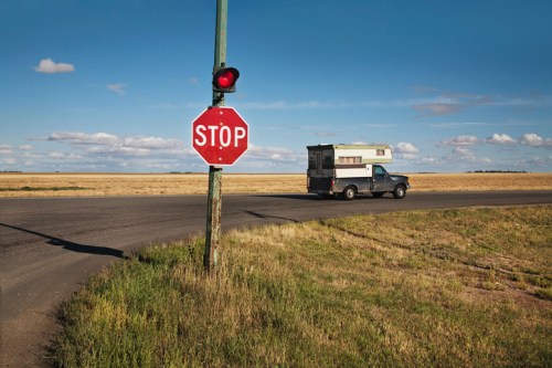 A truck with a camper drives past a stop sign on a rural road, with open fields and a blue sky in the background.