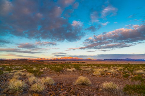 A vast desert landscape under a colorful sky, featuring mountains in the distance and scattered shrubs in the foreground.