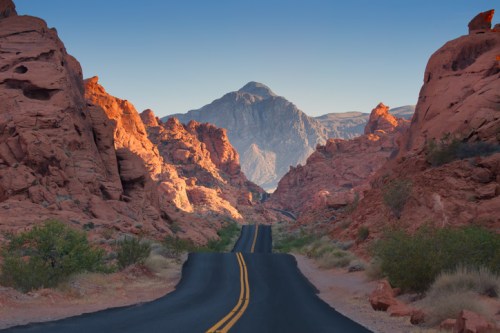 A winding road through red rock formations, leading towards a distant mountain under a clear blue sky.